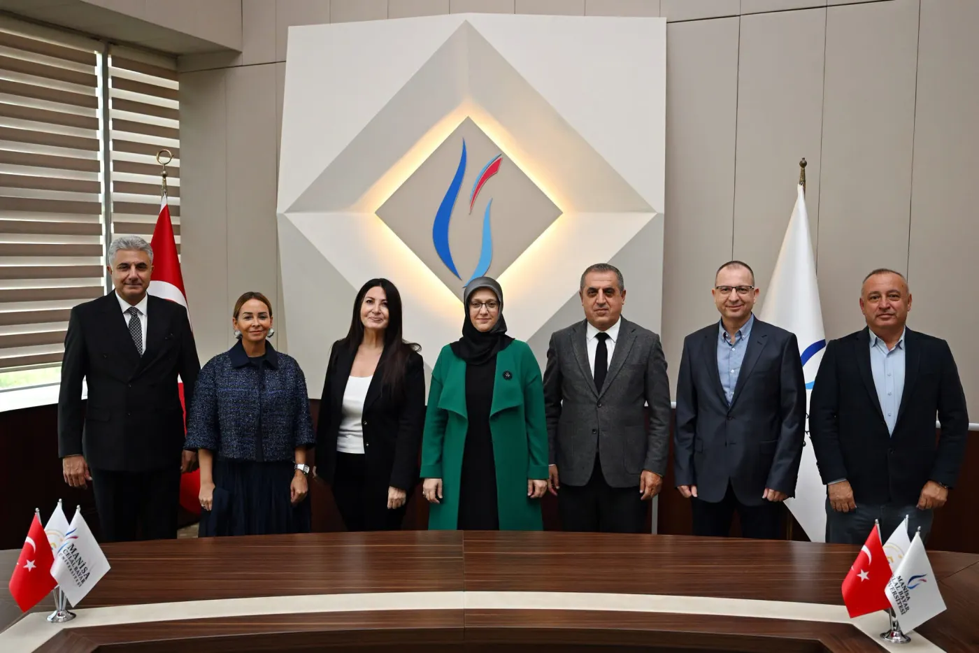 MCBÜ rektörlük toplantı salonunda, üniversite logosu ve bayraklar önünde toplantı masasının arkasında yan yana duran yedi kişilik heyet